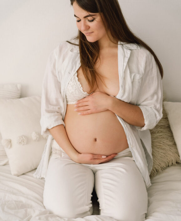 A beautiful pregnant woman, dressed in white clothes, is sitting in bed and holding her belly A beautiful pregnant woman, dressed in white clothes, is sitting in bed and holding her belly. Pregnancy concept.