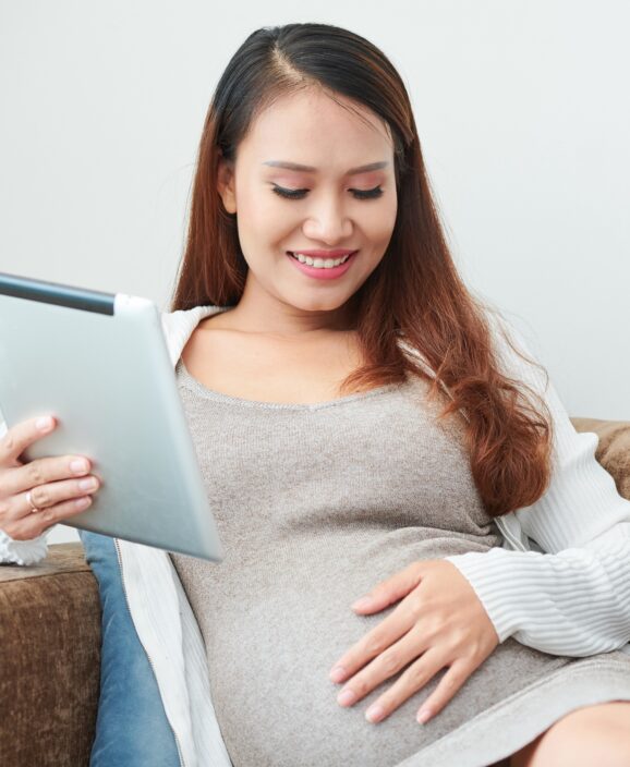 Pregnant woman resting on sofa Smiling young Asian pregnant woman resting on sofa with tablet computer in her hands and touching her belly